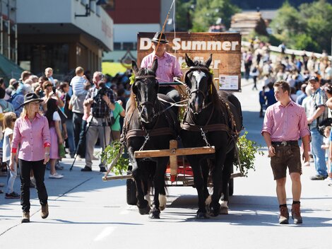 Ein Pferdewagen zieht durch eine belebte Straße. Viele Zuschauer in traditioneller Kleidung stehen entlang des Weges und beobachten die Parade.