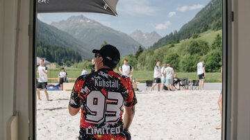 Ein Mann sitzt im Sand und schaut auf eine Gruppe von Menschen, die aktiv spielen. Im Hintergrund sind Berge und eine grüne Landschaft zu sehen.