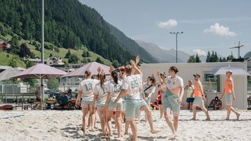 Eine Gruppe von Jugendlichen spielt im Sand und hat Spaß. Im Hintergrund sind Berge und Sonnenschirme zu sehen.