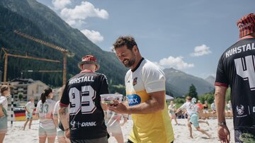 Eine Gruppe von Menschen in Sportkleidung auf einem sandigen Strand. Im Hintergrund sind Berge und ein blaues Himmel zu sehen.