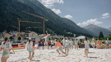 Eine Gruppe von jungen Sportlern spielt Beachvolleyball am Strand. Im Hintergrund sind Berge und ein belebtes Dorf zu sehen.