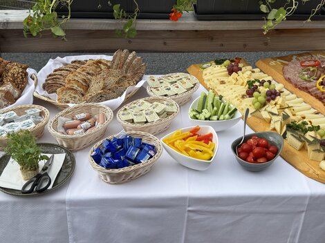A lavish buffet with various types of bread, cheese, cold cuts, and fresh vegetables. Colorful fruits and snacks complement the offerings on a table with a white tablecloth.