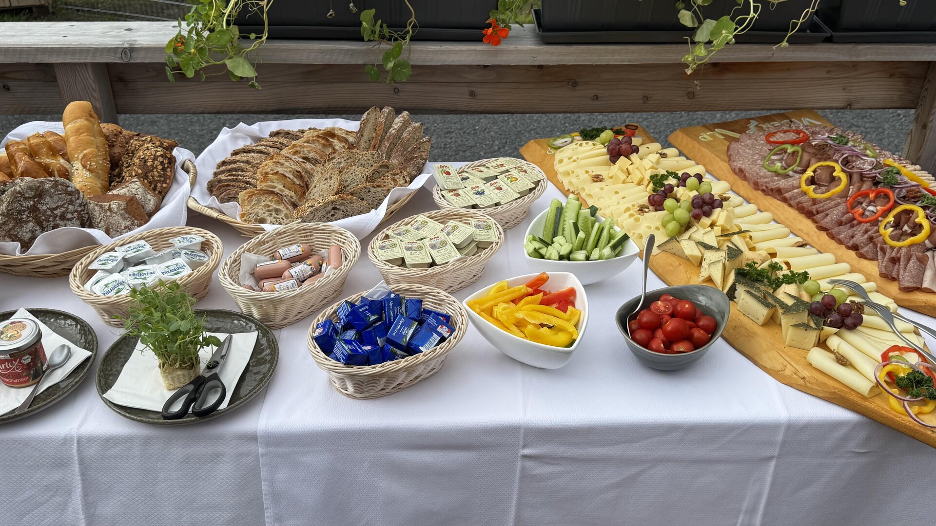 A lavish buffet with various types of bread, cheese, cold cuts, and fresh vegetables. Colorful fruits and snacks complement the offerings on a table with a white tablecloth.