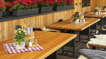 A cozy outdoor area with wooden tables and colorful flowers. Glasses and fresh plants are on the tables, creating an inviting atmosphere.