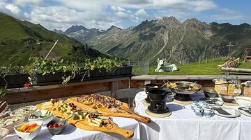 A set table with various dishes on a terrace in the mountains. In the background, green hills and a blue sky can be seen.