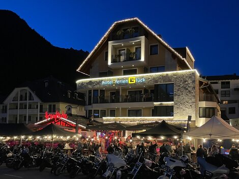 A lively hotel with a festively lit facade at dusk. In front of the hotel, there are many motorcycles and there is a bustling outdoor restaurant.