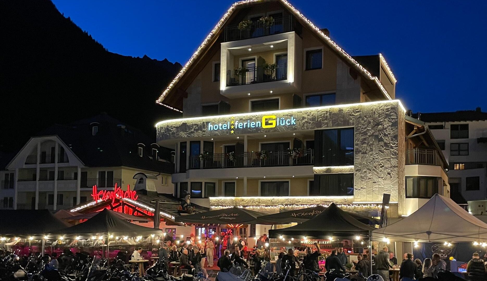 A lively hotel with a festively lit facade at dusk. In front of the hotel, there are many motorcycles and there is a bustling outdoor restaurant.