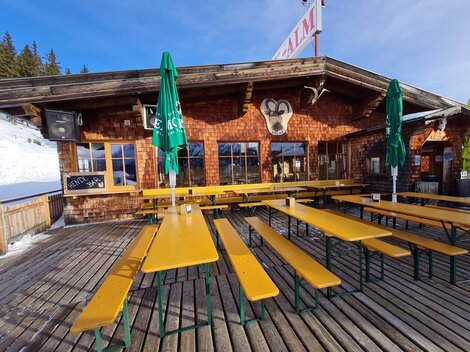 Eine rustikale Berghütte mit einer Holzterrasse und gelben Bänken. Es gibt grüne Sonnenschirme und verschneite Landschaft im Hintergrund.