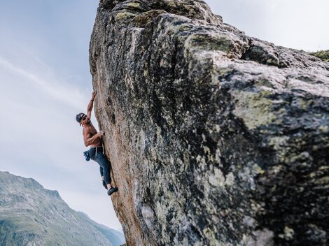 An athlete is climbing a steep rock face. The landscape in the background features mountains and a clear sky.