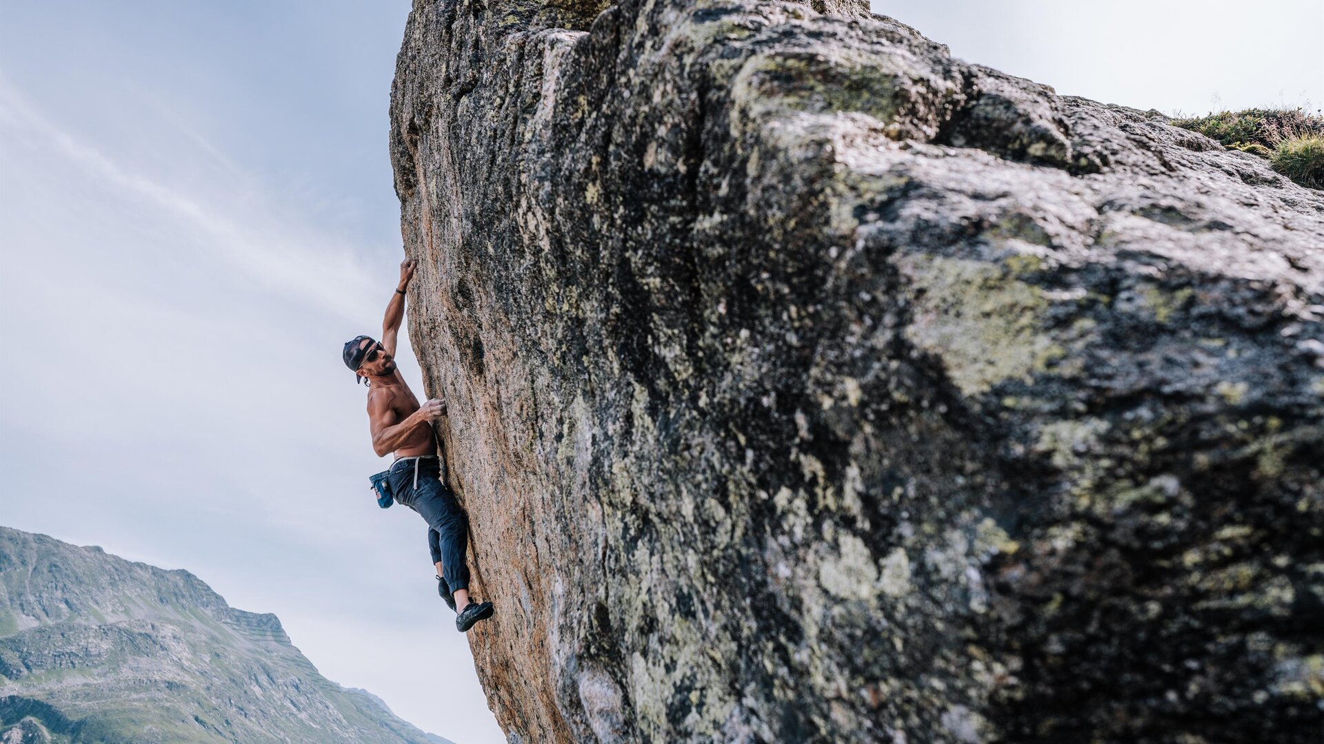 An athlete is climbing a steep rock face. The landscape in the background features mountains and a clear sky.