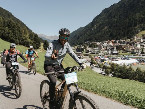 Eine Gruppe von Radfahrern auf einem malerischen Weg in den Bergen. Im Hintergrund ist ein kleiner Ort umgeben von grünen Wiesen und Bergen zu sehen.