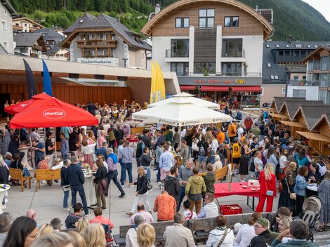 A bustling square with many people participating in a festival. Surrounded by stalls and buildings, the atmosphere radiates joy and camaraderie.