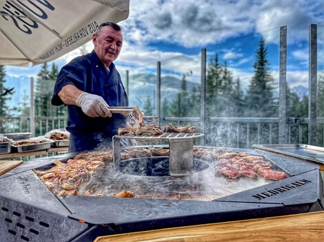 Ein Koch grillt Fleisch an einem runden Grill im Freien. Im Hintergrund sind Bäume und ein bewölkter Himmel zu sehen.