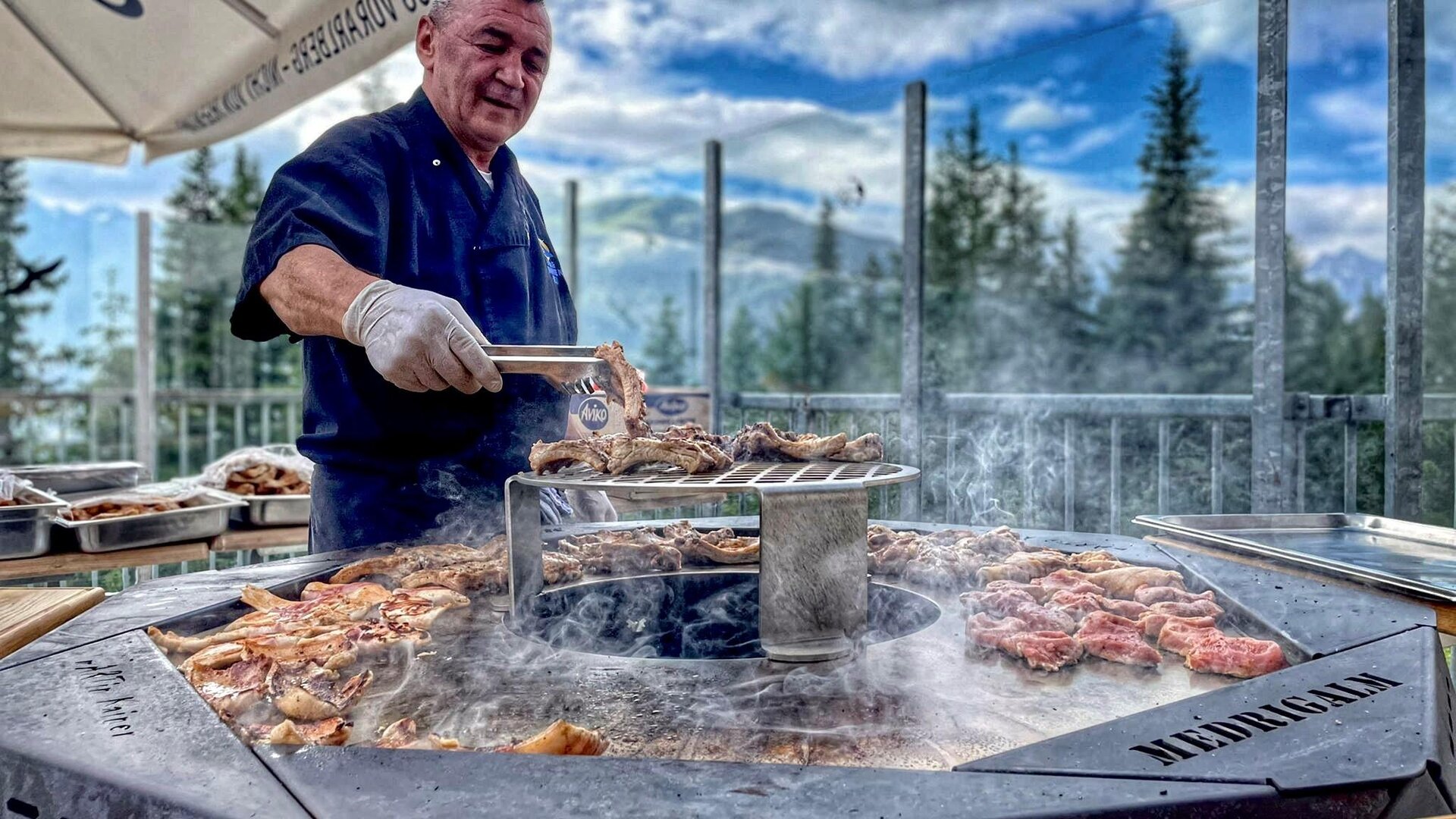 Ein Koch grillt Fleisch an einem runden Grill im Freien. Im Hintergrund sind Bäume und ein bewölkter Himmel zu sehen.