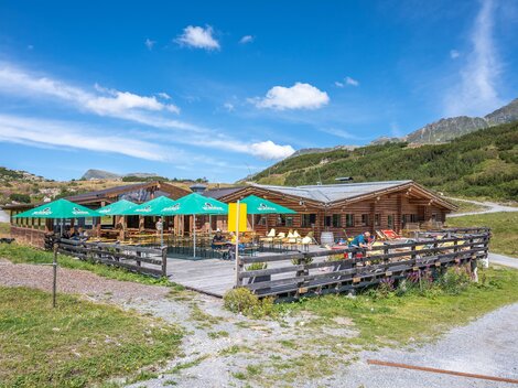 A cozy wooden house with a terrace and green sunshades. The surroundings are mountainous and green under a blue sky.