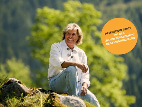 A smiling man is sitting on a rock in nature, surrounded by green trees. In the background, there is an event notice for an aftershow party.