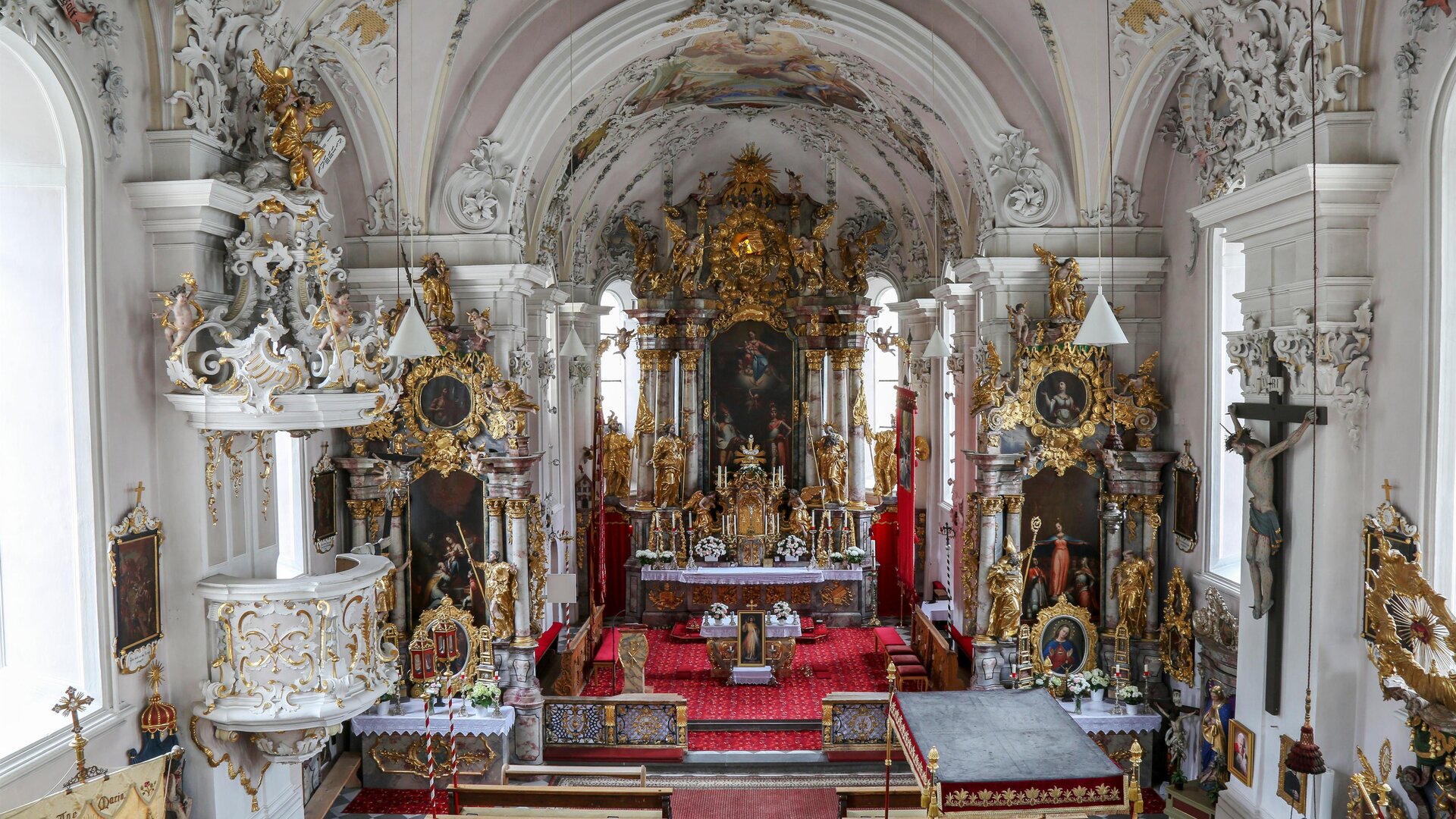 Eine prächtige Kirche mit reich verzierten Wänden und einem kunstvollen Altar. Der Innenraum ist lichtdurchflutet und strahlt eine spirituelle Atmosphäre aus.