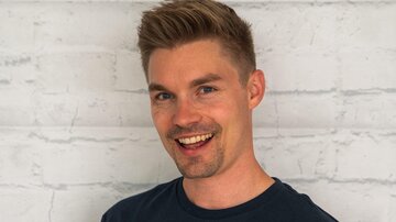 Ein junger Mann mit kurzem, blondem Haar lächelt freundlich. Er trägt ein schwarzes T-Shirt mit dem Aufdruck "ALPHATAURI".