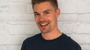 A young man with short, blond hair smiles kindly. He is wearing a black T-shirt with the print "ALPHATAURI".