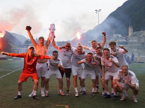 Eine Gruppe von Fußballspielern feiert auf dem Platz. Sie sind fröhlich und halten Fackeln in der Hand, während im Hintergrund eine malerische Berglandschaft zu sehen ist.