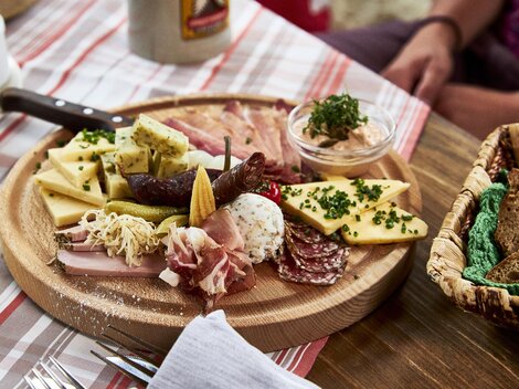 Eine rustikale Käse- und Wurstplatte auf einem Holzbrett. Dazu gibt es eingelegtes Gemüse und einen Dip im kleinen Schälchen.