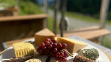 A plate with various types of cheese, grapes, and nuts. In the background, mountains and a clear sky can be seen.