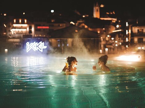 Two women are relaxing in the illuminated pool at night. The lights of the city are visible in the background.