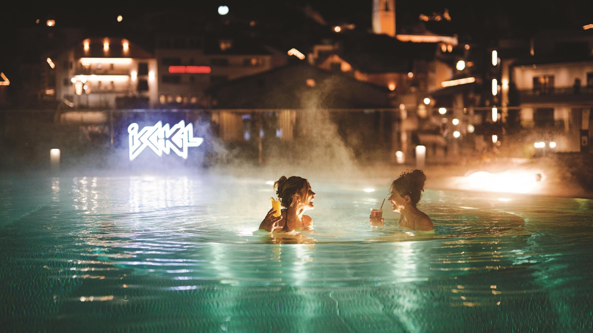 Two women are relaxing in the illuminated pool at night. The lights of the city are visible in the background.