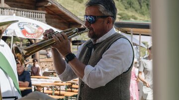 Ein Mann spielt Trompete in traditioneller Tracht. Im Hintergrund sind Tische und Bänke sowie grüne Berge zu sehen.