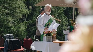 Ein Priester steht im Freien hinter einem Tisch mit Blumen und Geräten für eine Zeremonie. Im Hintergrund sind Stühle und Bäume zu sehen.