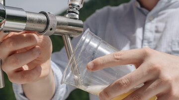 Ein junger Mann zapft ein Getränk aus einem Fass in ein Plastikbecher. Er trägt eine Sonnenbrille und einen Hut, während er in einer sonnigen Umgebung steht.