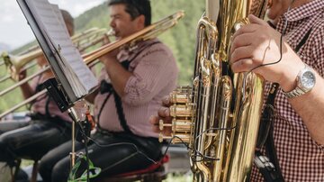 Eine Musikkapelle spielt im Freien, während sie traditionelle Trachten trägt. Im Vordergrund ist ein Musiker mit einer Tuba zu sehen.