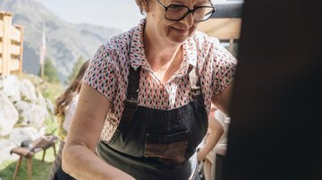 Eine Frau in einer Schürze arbeitet konzentriert an einem Tisch im Freien. Im Hintergrund sind Berge und ein klarer Himmel sichtbar.
