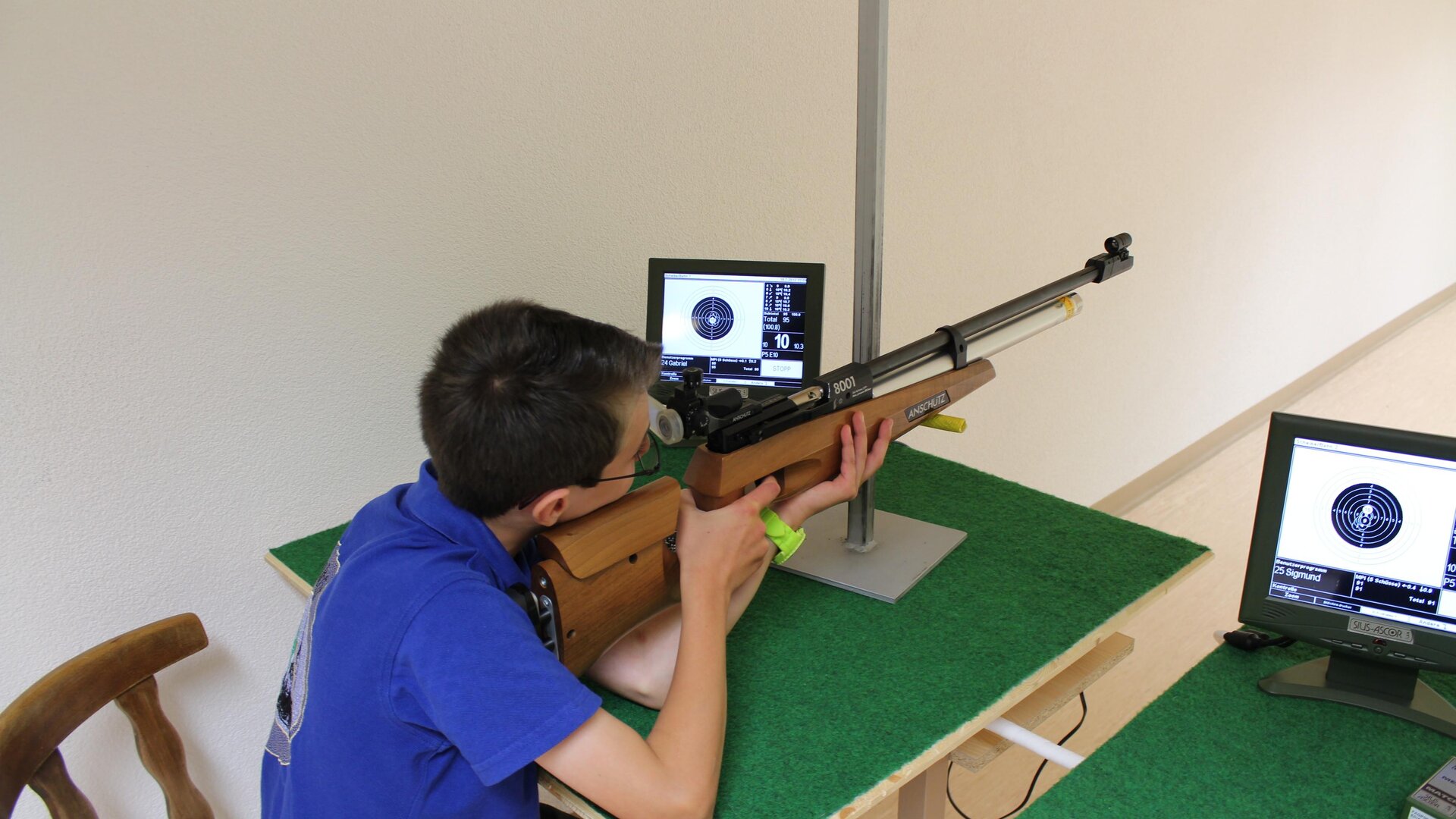Ein Junge schießt mit einem Luftgewehr auf ein Ziel. Vor ihm sind zwei Monitore, die seine Treffer anzeigen.