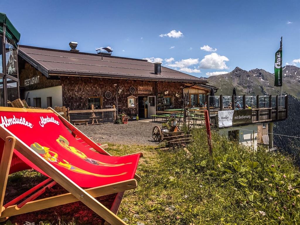A rustic mountain restaurant with a sun terrace. In the foreground is a red deck chair on a green meadow.