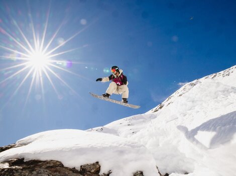 A snowboarder jumps over a snow-covered hill under a bright blue sky. The sun shines brightly, creating a dynamic atmosphere.