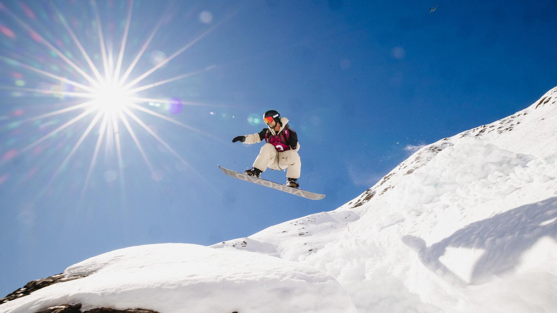 A snowboarder jumps over a snow-covered hill under a bright blue sky. The sun shines brightly, creating a dynamic atmosphere.