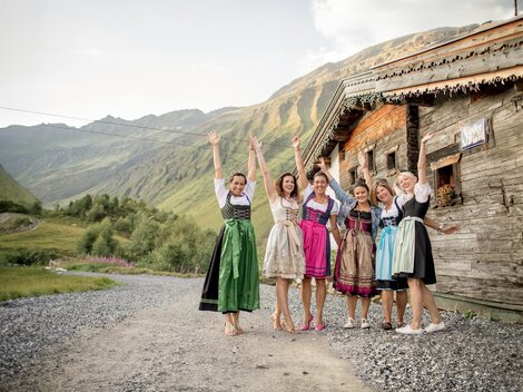 Sechs Frauen in traditioneller Tracht stehen fröhlich vor einem alten Holzhaus in den Bergen. Die Landschaft ist grün und hügelig mit einer beeindruckenden Bergkulisse.