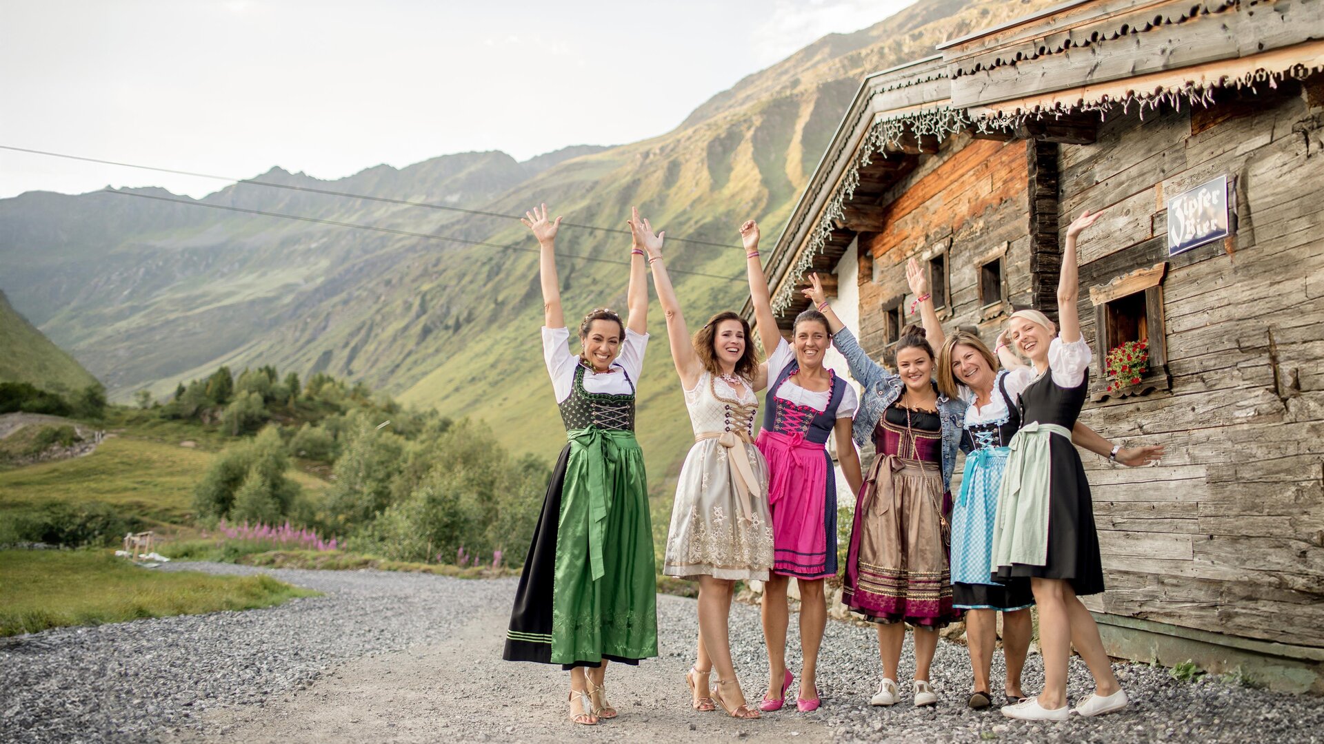 Six women in traditional attire stand joyfully in front of an old wooden house in the mountains. The landscape is green and hilly with an impressive mountain backdrop.
