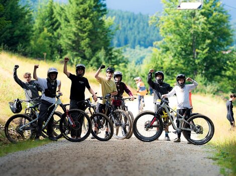 A group of cyclists stands on a gravel path in nature. They excitedly raise their fists and wear helmets, while other people are on the move in the background.
