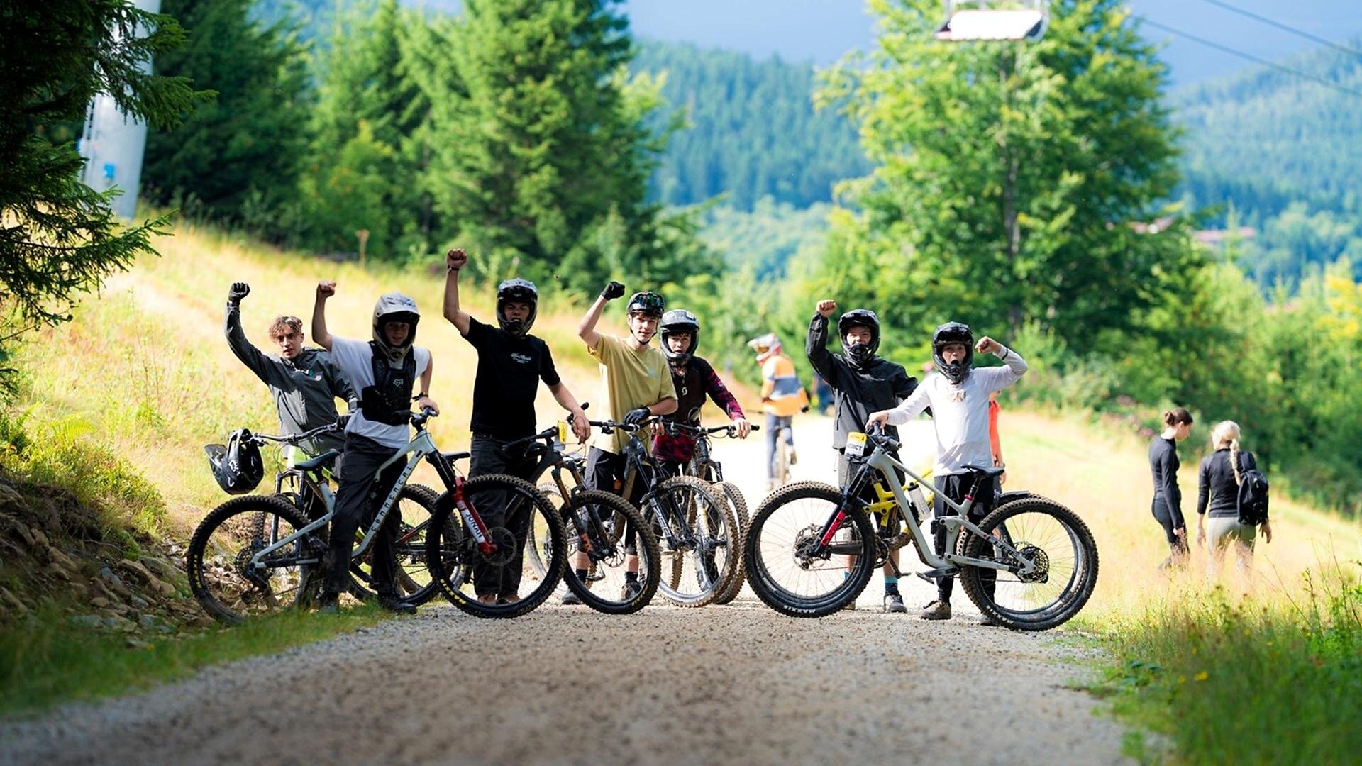 A group of cyclists stands on a gravel path in nature. They excitedly raise their fists and wear helmets, while other people are on the move in the background.