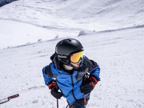 Ein Junge im blauen Skianzug trägt einen Helm und eine Skibrille. Er steht auf einem schneebedeckten Hang und schaut nach oben.