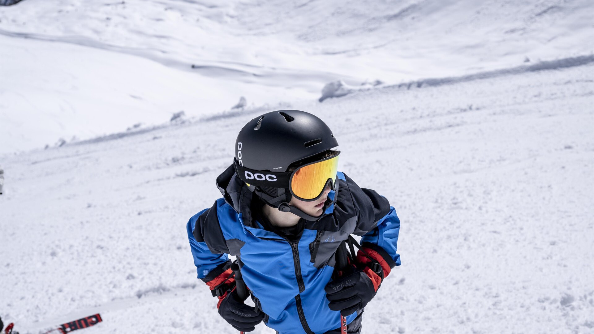 Ein Junge im blauen Skianzug trägt einen Helm und eine Skibrille. Er steht auf einem schneebedeckten Hang und schaut nach oben.