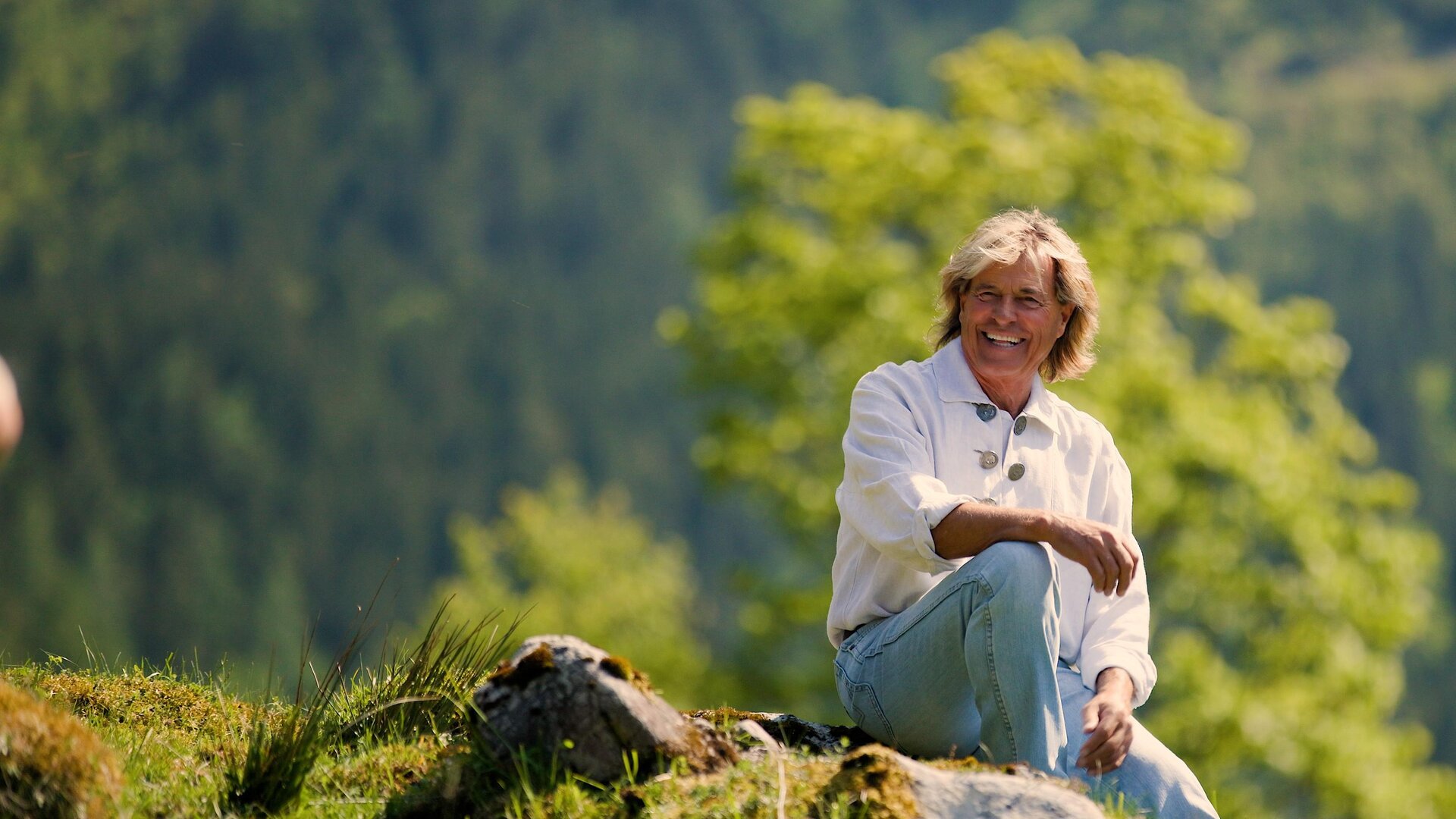 A man is sitting relaxed on a meadow with trees in the background. The surroundings are natural and peaceful.
