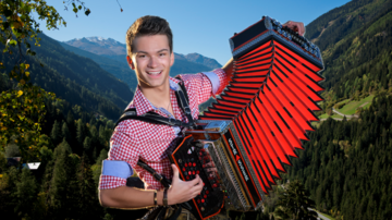 A young man is playing an accordion in the mountains. The landscape is green with snow-capped peaks in the background.