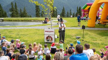 A large group of children is sitting on the grass watching a performer. In the background, an inflatable bounce house and beautiful mountains can be seen.