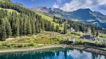 Eine schöne Berglandschaft mit grünen Wäldern und einem klaren See. Im Hintergrund sind majestätische Berge und einige Gebäude sichtbar.