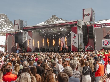 A large music festival in the mountains with an impressive stage. Many people enjoy the performance under clear skies.