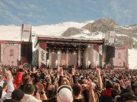 A large music festival in the mountains with many people dancing to the show. In the background, snow-covered mountains and a stage with lights can be seen.