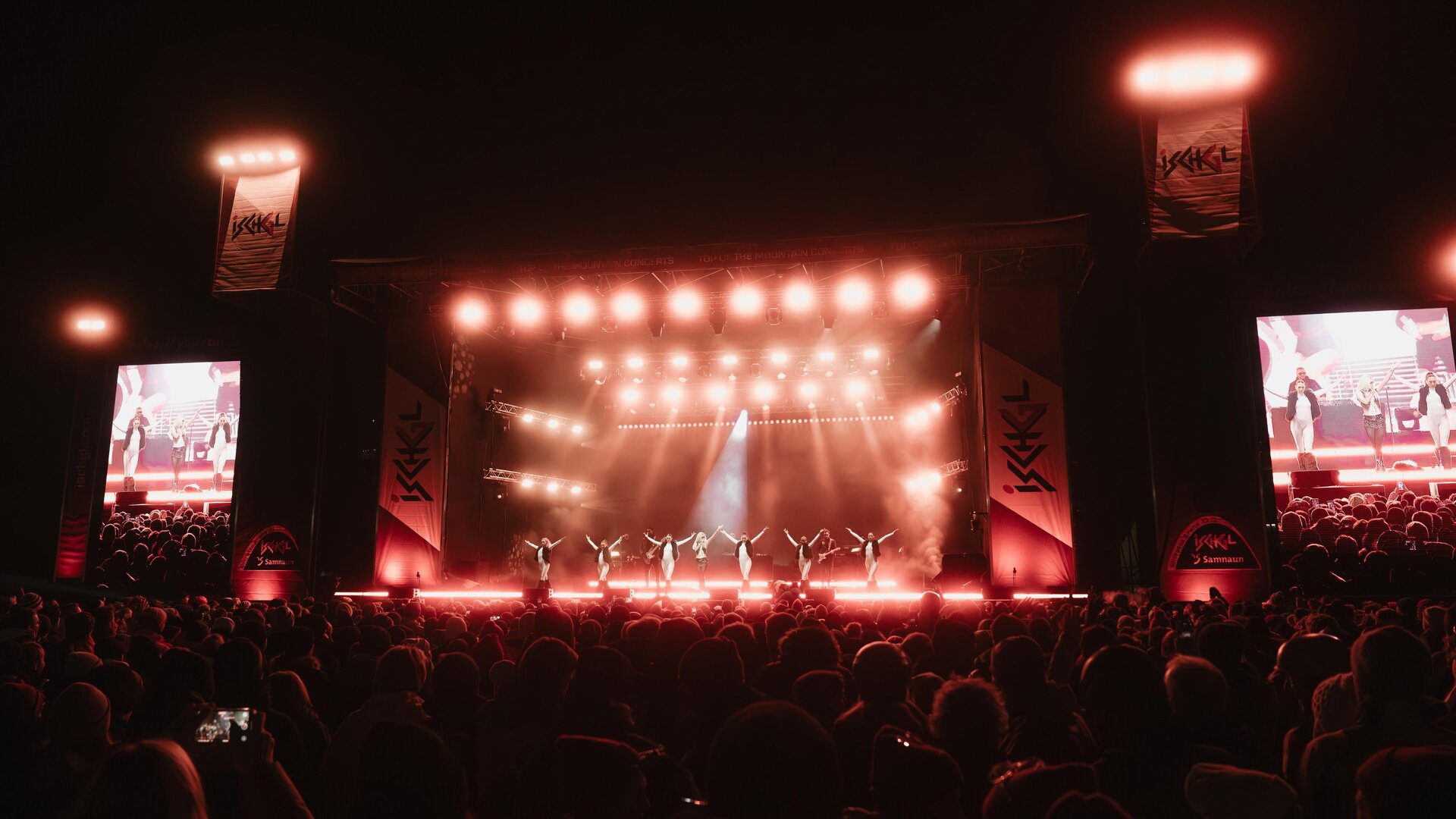 A large crowd is watching a stage performance at night. The stage is brightly lit, with red lights and several screens.